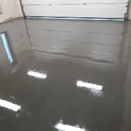 Gładka, szara posadzka w garażu z otwartą, białą bramą segmentową i widocznymi refleksami światła.