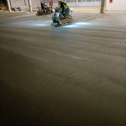 Wylewki.pl - Wyrównywanie wylewki w hali przemysłowej za pomocą zacieraczki mechanicznej, widoczne ślady po pracy maszyny i dwóch pracowników w tle.