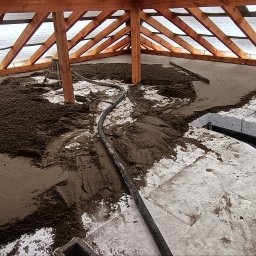 Wylewki.pl - Wnętrze budynku w trakcie wylewania posadzki: ciemna masa wylewki częściowo rozprowadzona na białej izolacji podłogowej, widoczna rura doprowadzająca materiał, drewniana konstrukcja dachu...
