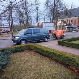 Wylewki.pl - Szary van ciągnie pomarańczową maszynę do wylewania posadzek po mokrej ulicy w miejskiej scenerii z kościołem w tle.