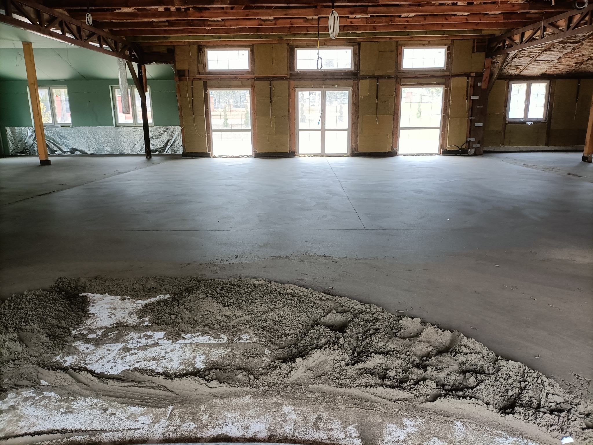 Wnętrze budynku w trakcie wylewania posadzki: widoczny jastrych w stanie surowym oraz fragment gładkiej, wylanej powierzchni. Ściany ocieplone wełną mineralną, drewniana konstrukcja dachu.