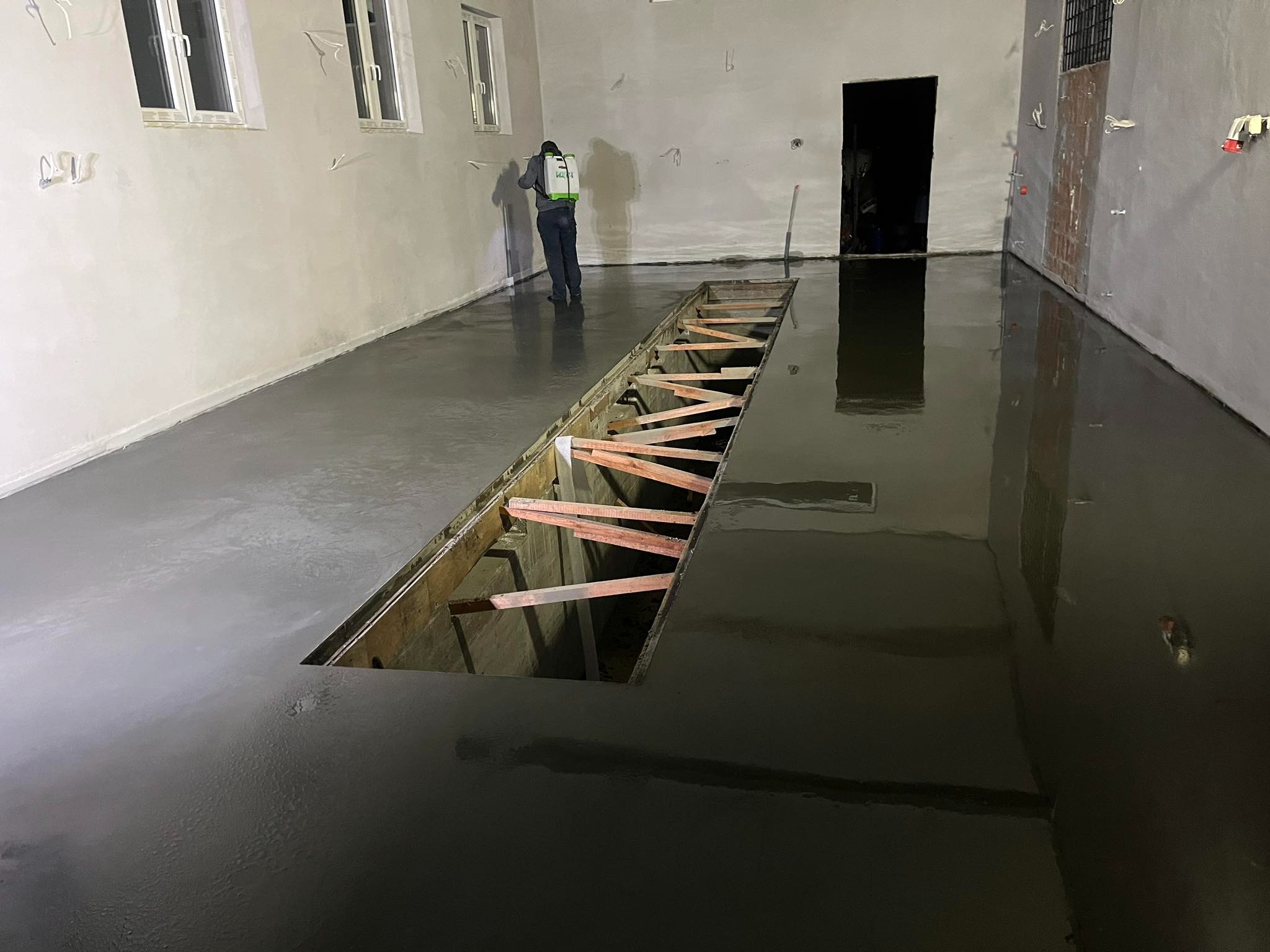 Aplikacja żywicy epoksydowej na posadzce przemysłowej w hali, widoczny otwór w podłodze z drewnianym szalunkiem i pracownik w kombinezonie ochronnym rozpylający preparat.
