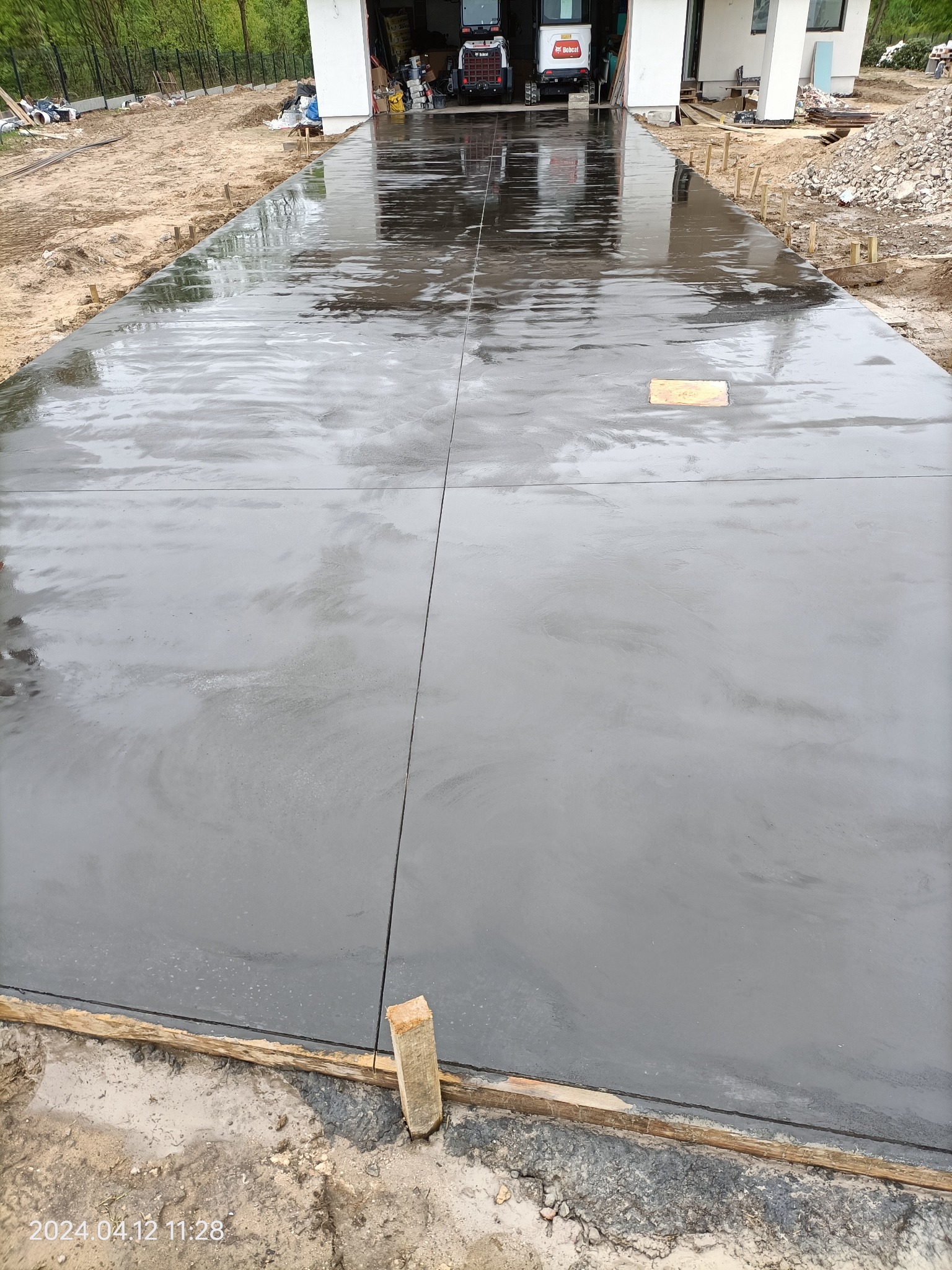Świeżo wylana, mokra betonowa posadzka na podjeździe, z widocznymi dylatacjami i deskowaniem obrzeży. W tle budynek z otwartym garażem i zaparkowanymi maszynami budowlanymi.