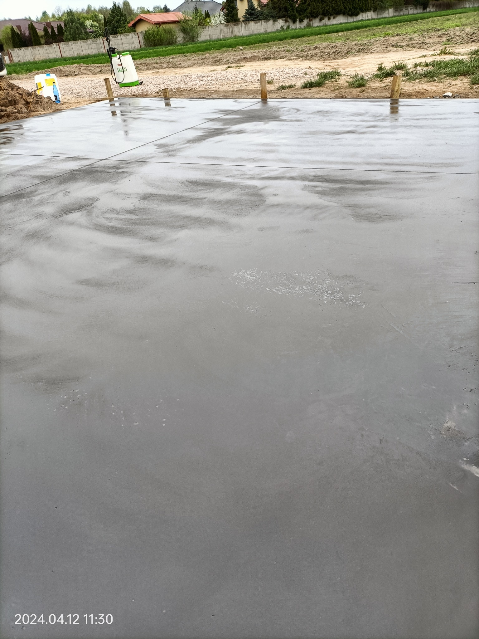 Świeżo wylana, gładka, szara posadzka z widocznymi drewnianymi kołkami i opryskiwaczem ciśnieniowym w tle, na zewnątrz w słoneczny dzień.