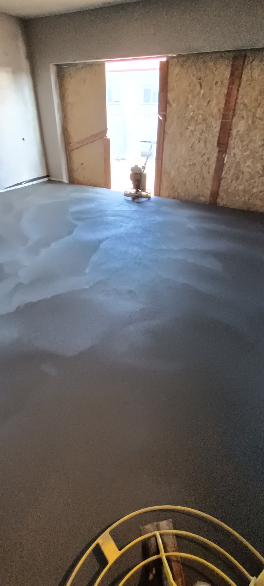 Wygładzona, szara posadzka w surowym wnętrzu, z maszyną do zacierania widoczną w tle, przy otwartych drzwiach prowadzących na zewnątrz.