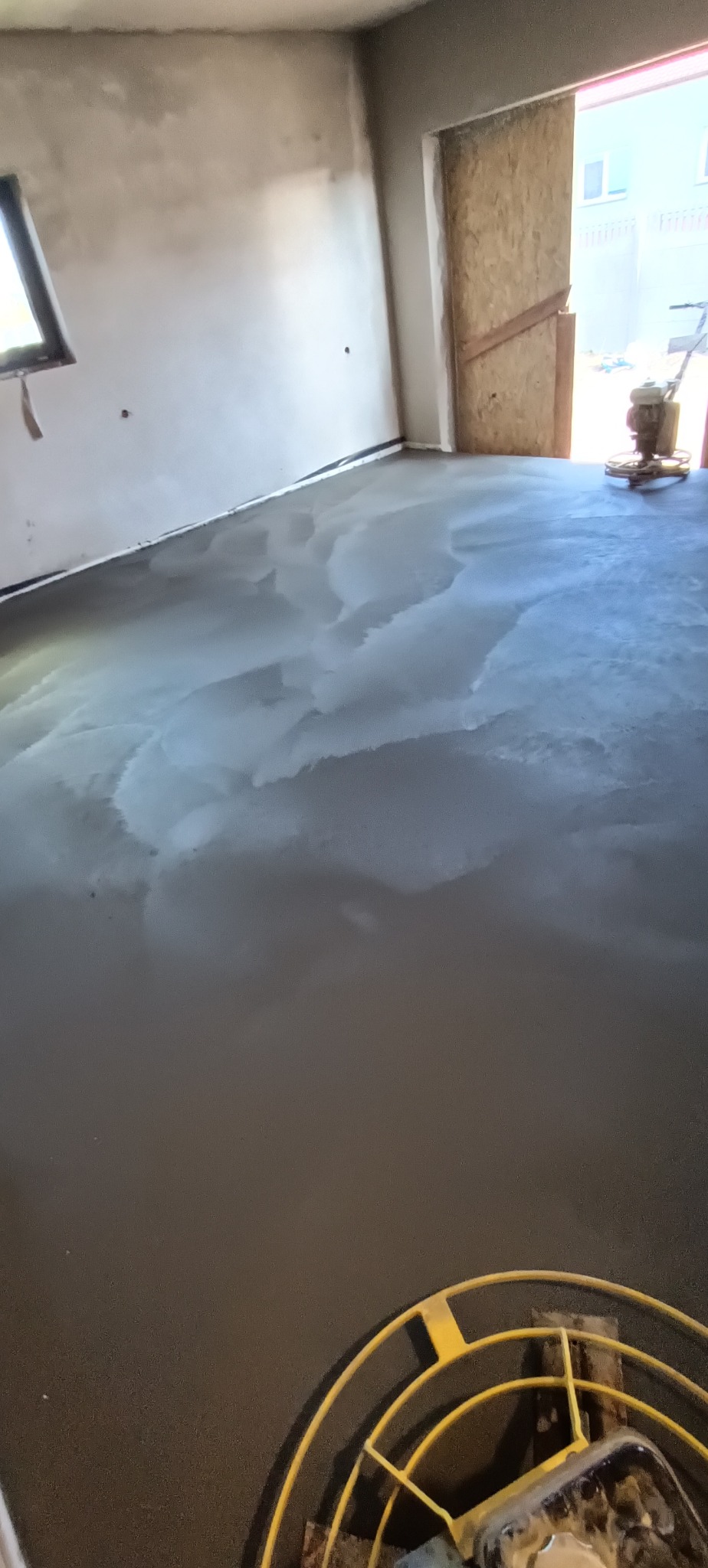 Świeżo wylana, mokra posadzka w surowym pomieszczeniu z widoczną maszyną do zacierania na drugim planie i fragmentem maszyny na pierwszym planie.