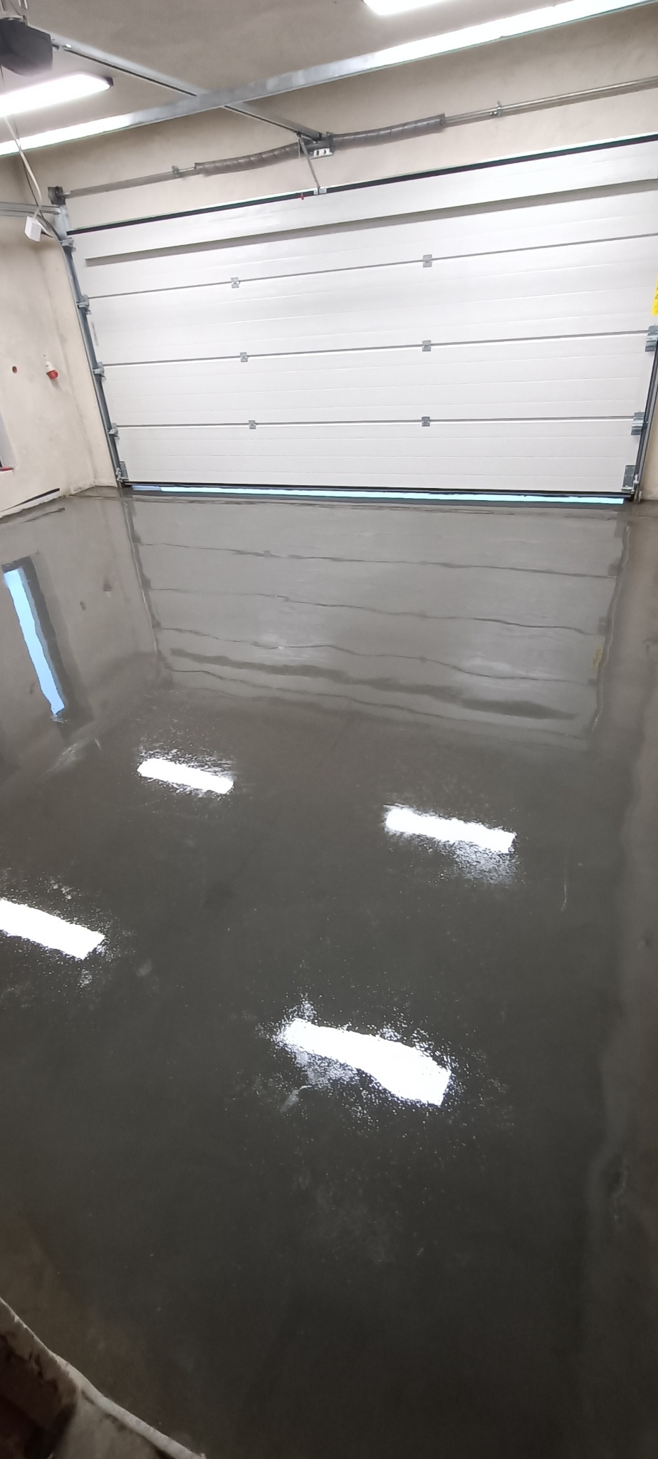 Gładka, szara posadzka w garażu z otwartą, białą bramą segmentową i widocznymi refleksami światła.