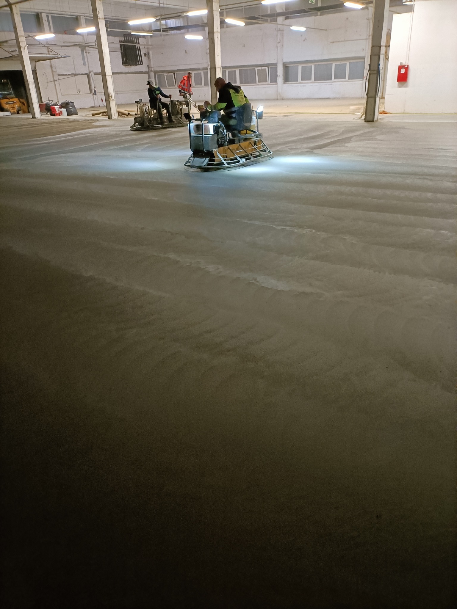 Wyrównywanie wylewki w hali przemysłowej za pomocą zacieraczki mechanicznej, widoczne ślady po pracy maszyny i dwóch pracowników w tle.