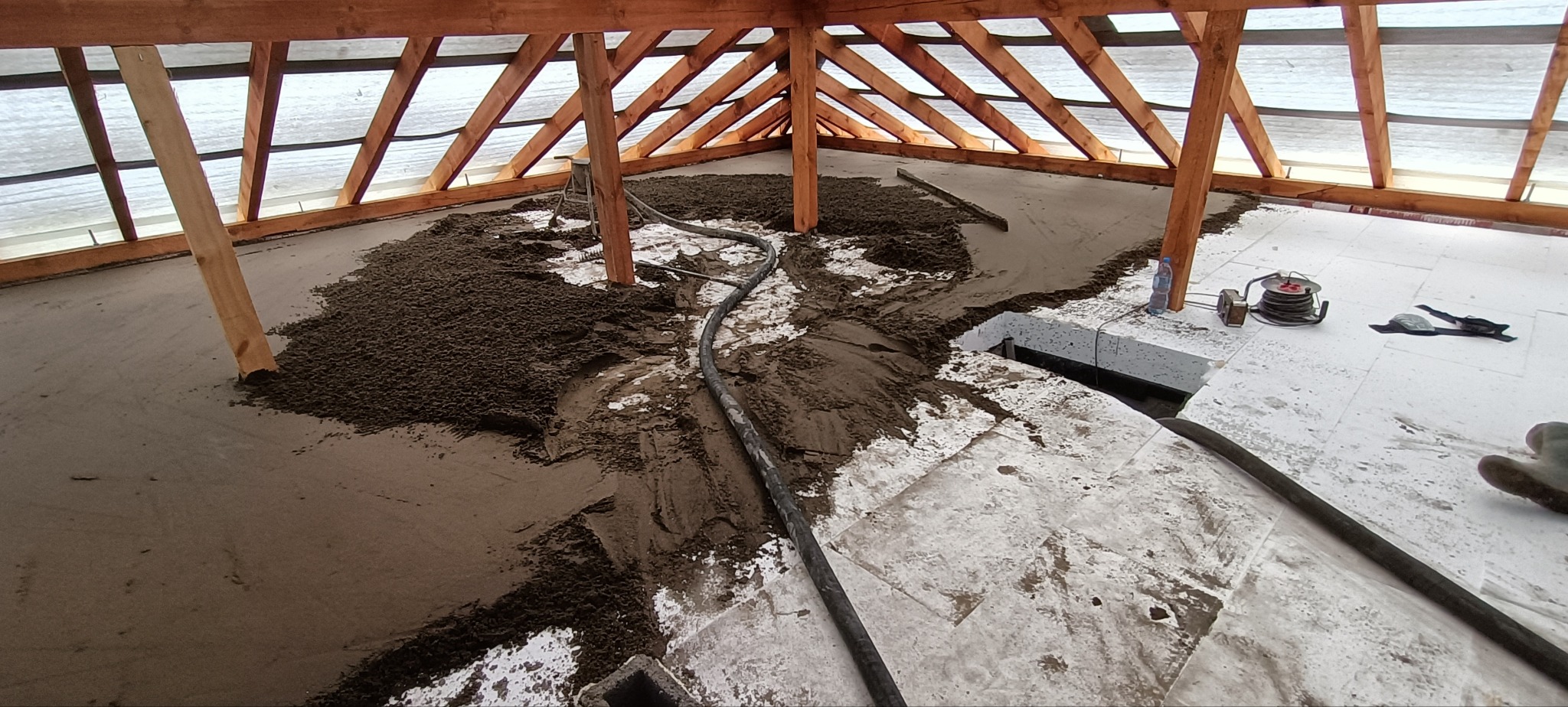 Wnętrze budynku w trakcie wylewania posadzki: ciemna masa wylewki częściowo rozprowadzona na białej izolacji podłogowej, widoczna rura doprowadzająca materiał, drewniana konstrukcja dachu...