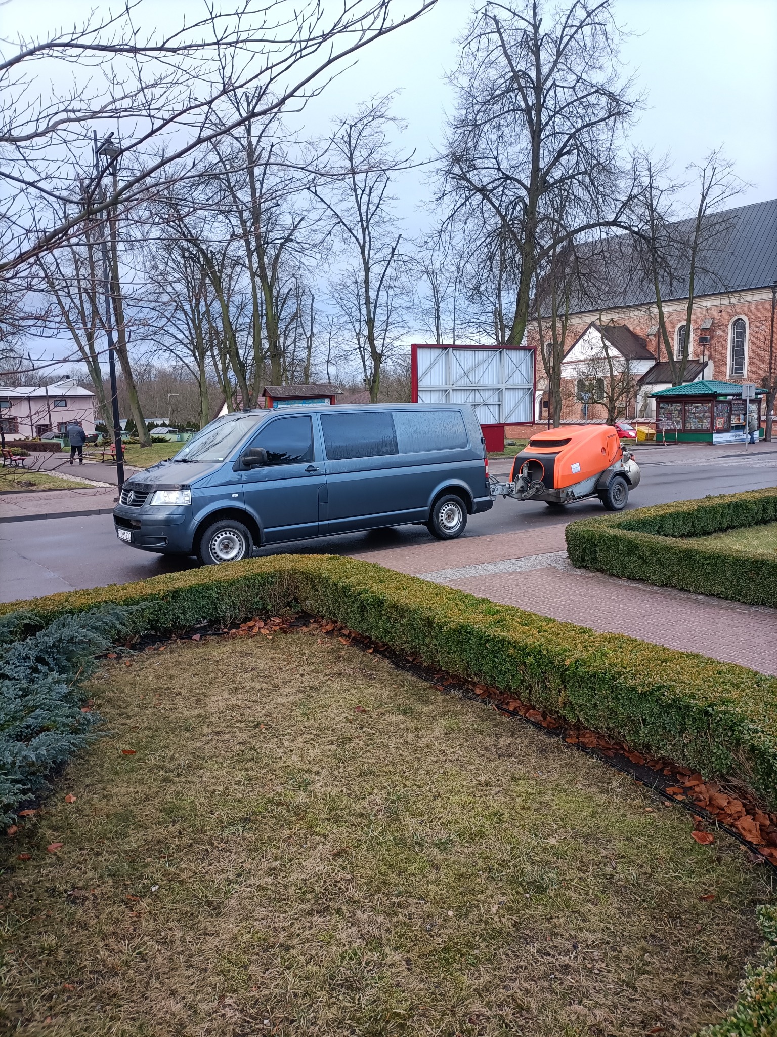 Szary van ciągnie pomarańczową maszynę do wylewania posadzek po mokrej ulicy w miejskiej scenerii z kościołem w tle.