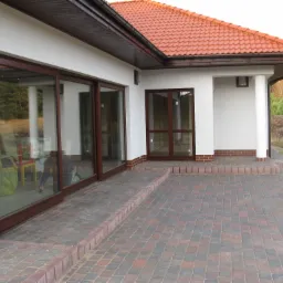 Nowoczesny dom murowany z czerwonym dachem, dużymi przesuwnymi oknami w brązowych ramach i brukowanym tarasem z kostki w odcieniach czerwieni i szarości.