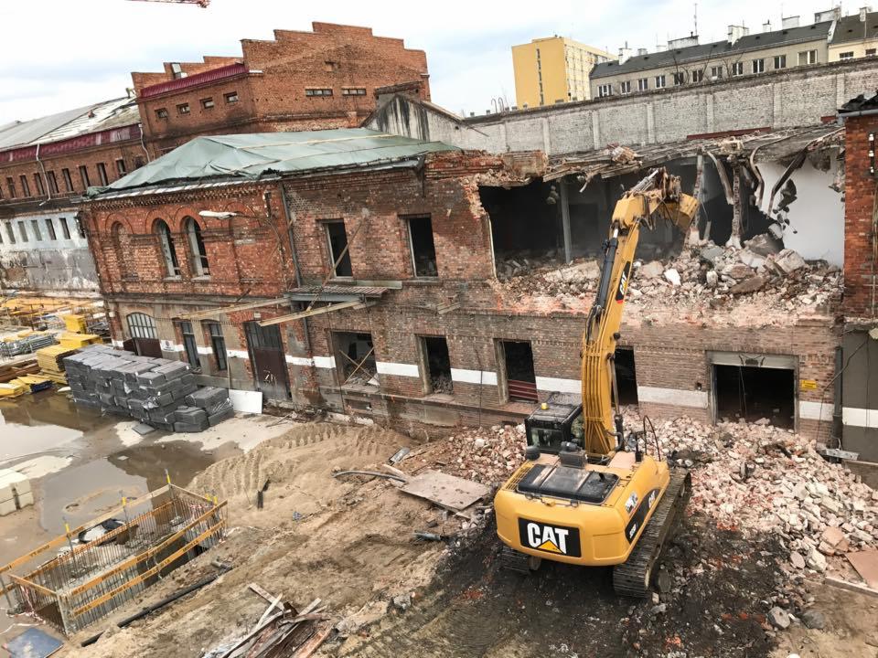 Żółta koparka CAT w trakcie prac rozbiórkowych budynku z czerwonej cegły w Warszawie. Częściowo zburzona konstrukcja, gruz i odsłonięte wnętrze budynku.