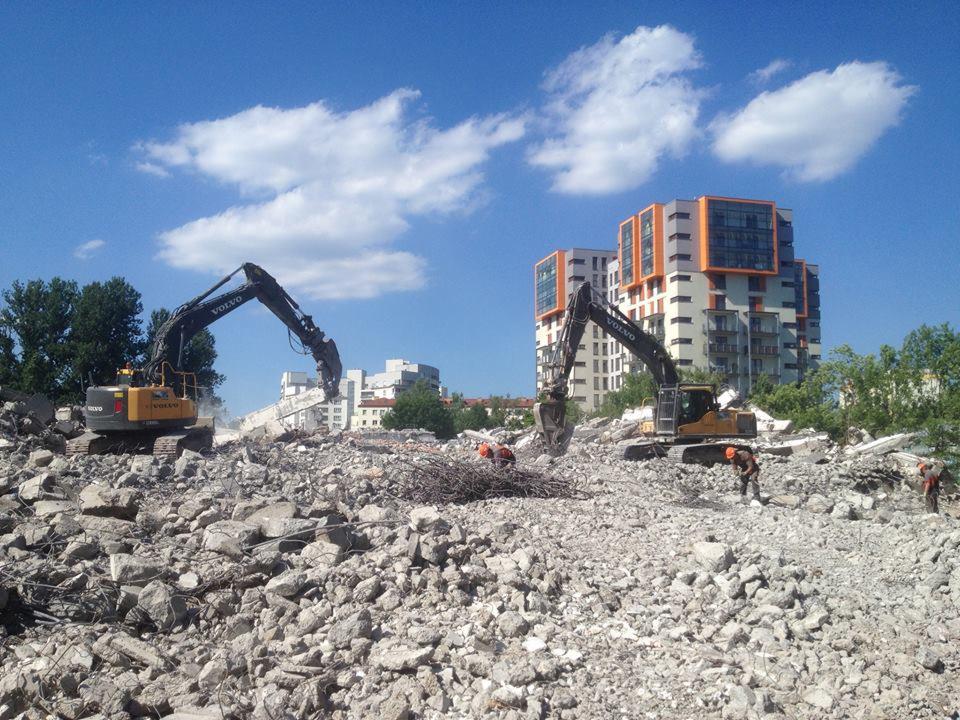 Dwa ciężkie, żółte koparki Volvo z hydraulicznymi nożycami kruszą betonowe gruzy na placu budowy; w tle widoczny wielopiętrowy budynek mieszkalny z pomarańczowymi akcentami i błękitne niebo...