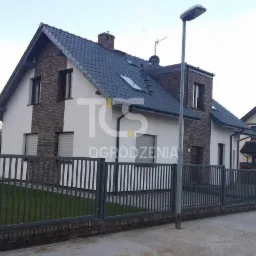 Nowoczesne ogrodzenie metalowe w kolorze grafitowym, z niską podmurówką z cegły, na tle domu z szarą dachówką i elementami ceglanymi. Słup oświetleniowy uliczny.