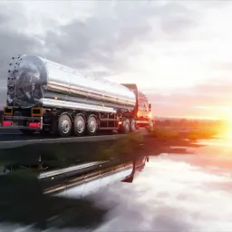 Cysterna transportowa w ruchu, odbicie w mokrej nawierzchni drogi, zachodzące słońce w tle.