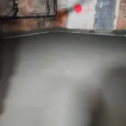 Świeżo wylana, gładka posadzka w surowym pomieszczeniu z widocznymi rurami instalacyjnymi na ścianie i śladami po remoncie.