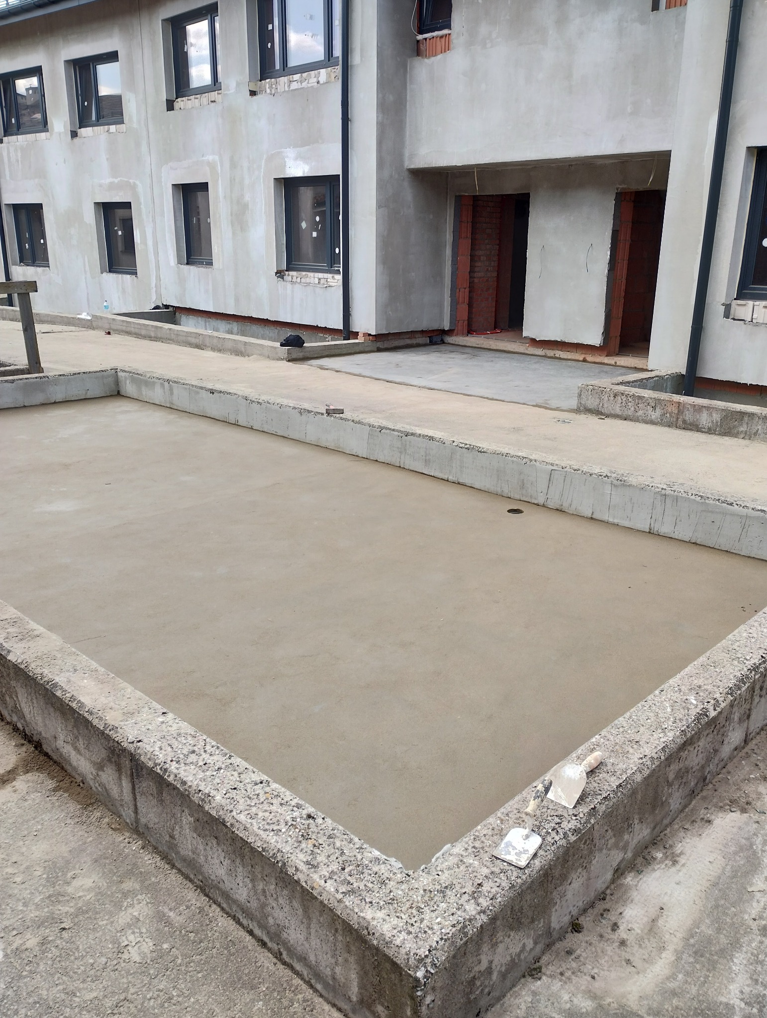 Wylana, gładka posadzka w betonowej ramie, z widocznymi narzędziami (szpachelki) na krawędzi, budynek w tle w stanie surowym.