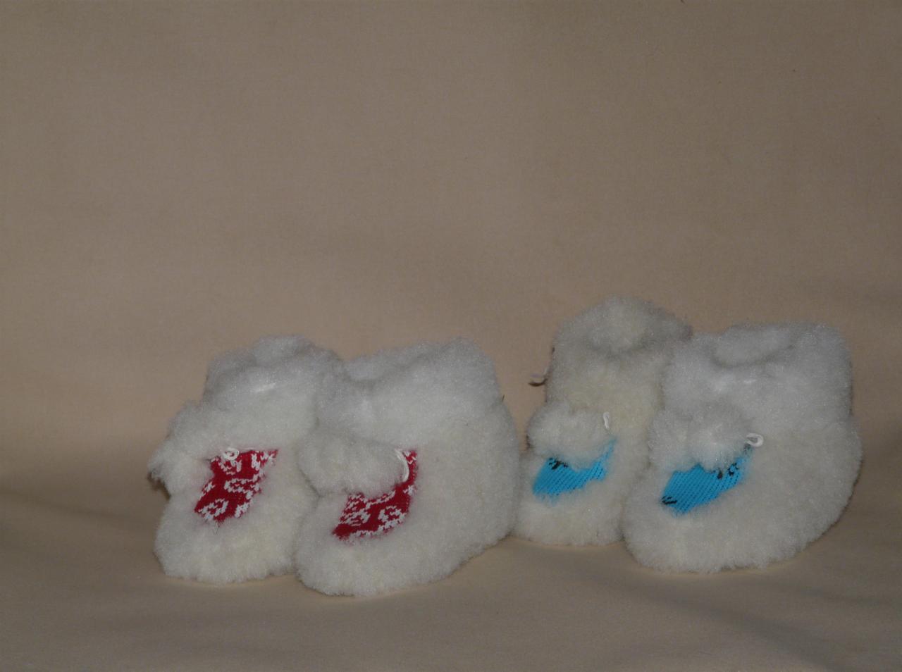 Dwie pary białych, wełnianych, ręcznie robionych bucików dla niemowląt z kolorowymi, haftowanymi aplikacjami na beżowym tle.