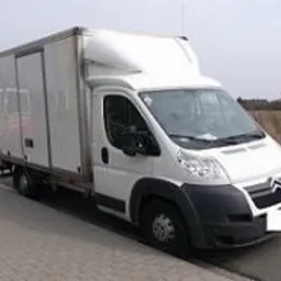 Biały samochód dostawczy z zabudową kontenerową i windą załadowczą, zaparkowany na poboczu drogi.