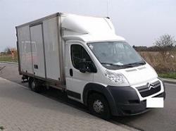 Biały samochód dostawczy z zabudową kontenerową i windą załadowczą, zaparkowany na poboczu drogi.