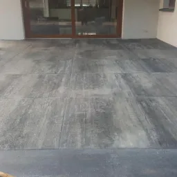 Nowoczesny taras wyłożony dużymi, szarymi płytami z widoczną fakturą drewna, przylegający do budynku z przesuwnymi drzwiami tarasowymi.