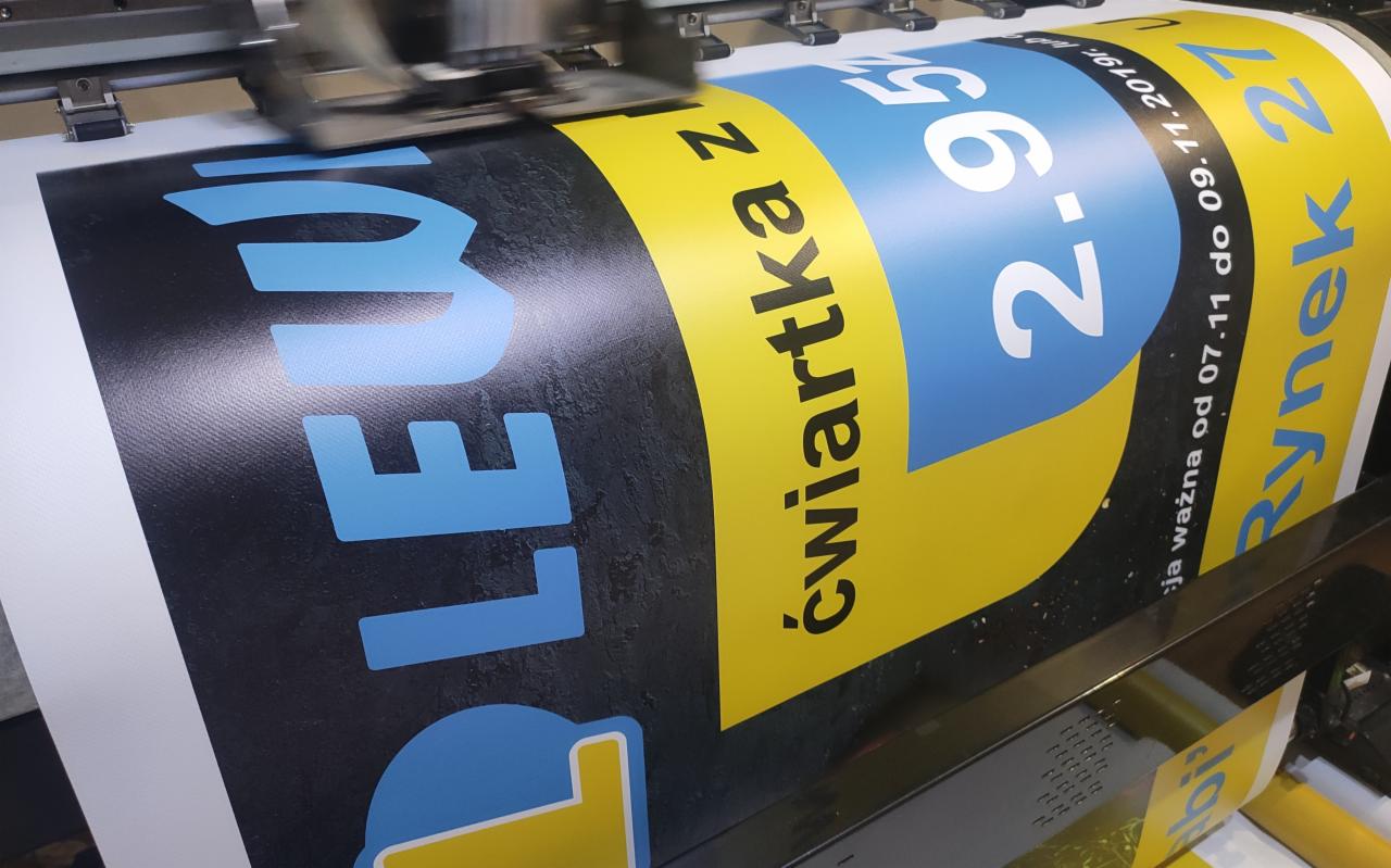 Druk wielkoformatowy: Proces drukowania plakatu promocyjnego z napisem 'ćwiartka z LEW' oraz informacją o cenie i dacie ważności oferty. Widoczna głowica drukująca w trakcie pracy.