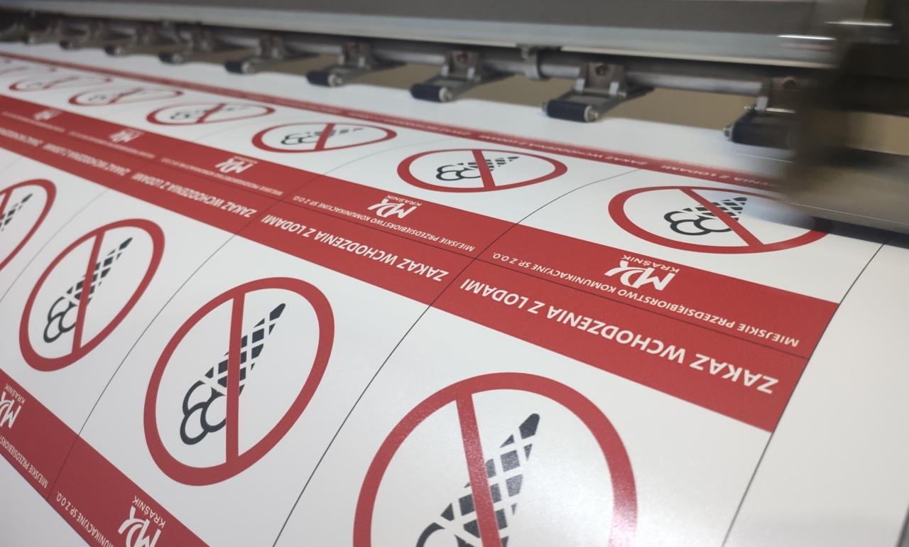 Drukarka przemysłowa drukuje serię okrągłych, samoprzylepnych etykiet z symbolem zakazu spożywania lodów, z czerwonym obramowaniem i napisem 'ZAKAZ WCHODZENIA Z LODAMI' oraz logo 'MD Krasnik'.