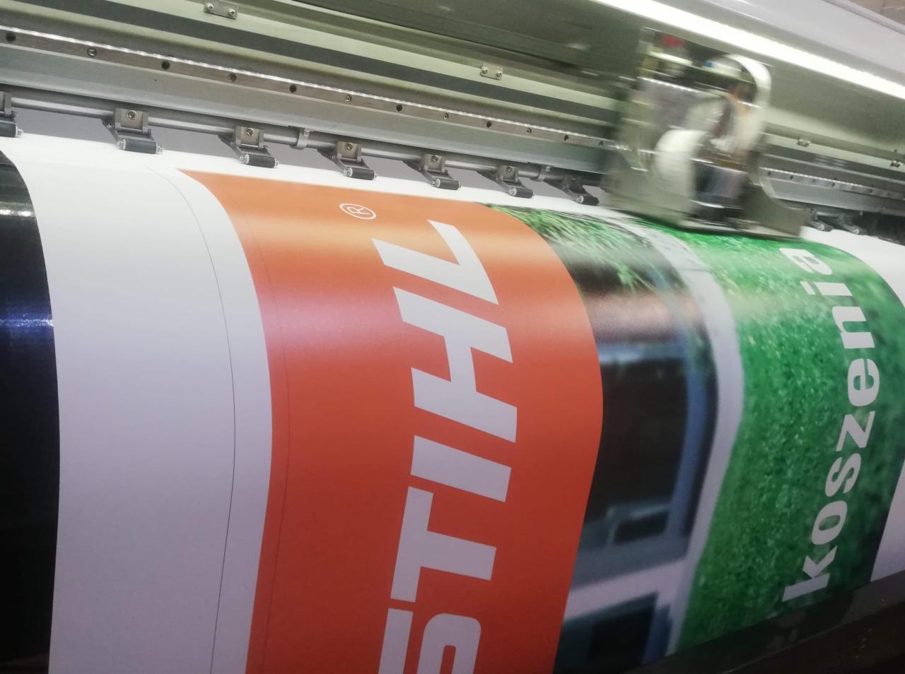 Drukarka wielkoformatowa w trakcie drukowania baneru reklamowego z logo STIHL i hasłem 'koszenia'