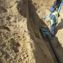 Wąski wykop w piaszczystej ziemi z widoczną maszyną do przeciskania rur i fragmentem rury, pracownik w odblaskowej kamizelce nadzoruje proces.