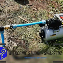 Szara hydrofornia Steinberg z podłączonymi niebieskimi rurami na trawiastym terenie, żółty wąż doprowadzający wodę, widoczny fragment reklamy Wiercenie Studni - Swarzędz.