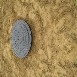 Szara, okrągła pokrywa włazu w ziemi, otoczona świeżo naruszoną glebą i fragmentami trawy, widoczne ślady stóp w ziemi.