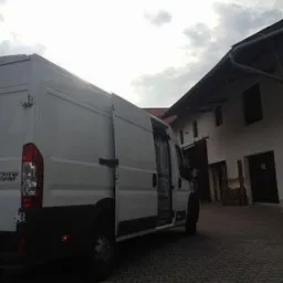 Biały furgon dostawczy Fiat Ducato Maxi z otwartymi tylnymi drzwiami, zaparkowany na brukowanym podwórzu przed budynkiem z ciemnymi okiennicami i dachem spadzistym.