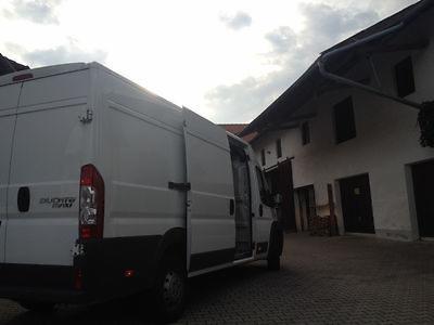 Biały furgon dostawczy Fiat Ducato Maxi z otwartymi tylnymi drzwiami, zaparkowany na brukowanym podwórzu przed budynkiem z ciemnymi okiennicami i dachem spadzistym.