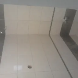 Wykończona łazienka z jasnymi płytkami i szarymi pasami na ścianach. Widoczny fragment podłogi z otworem. Górne rogi pomieszczenia lekko zaokrąglone.