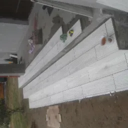 Układanie jasnych płytek na schodach wejściowych, widoczny kubek z napojem i narzędzia glazurnicze. Prace w toku, częściowo ukończone schody.