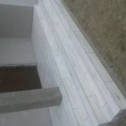 Ułożona jasna okładzina z płytek imitujących drewno na schodach wejściowych, widok z góry pod kątem, z fragmentem elewacji budynku w tle.