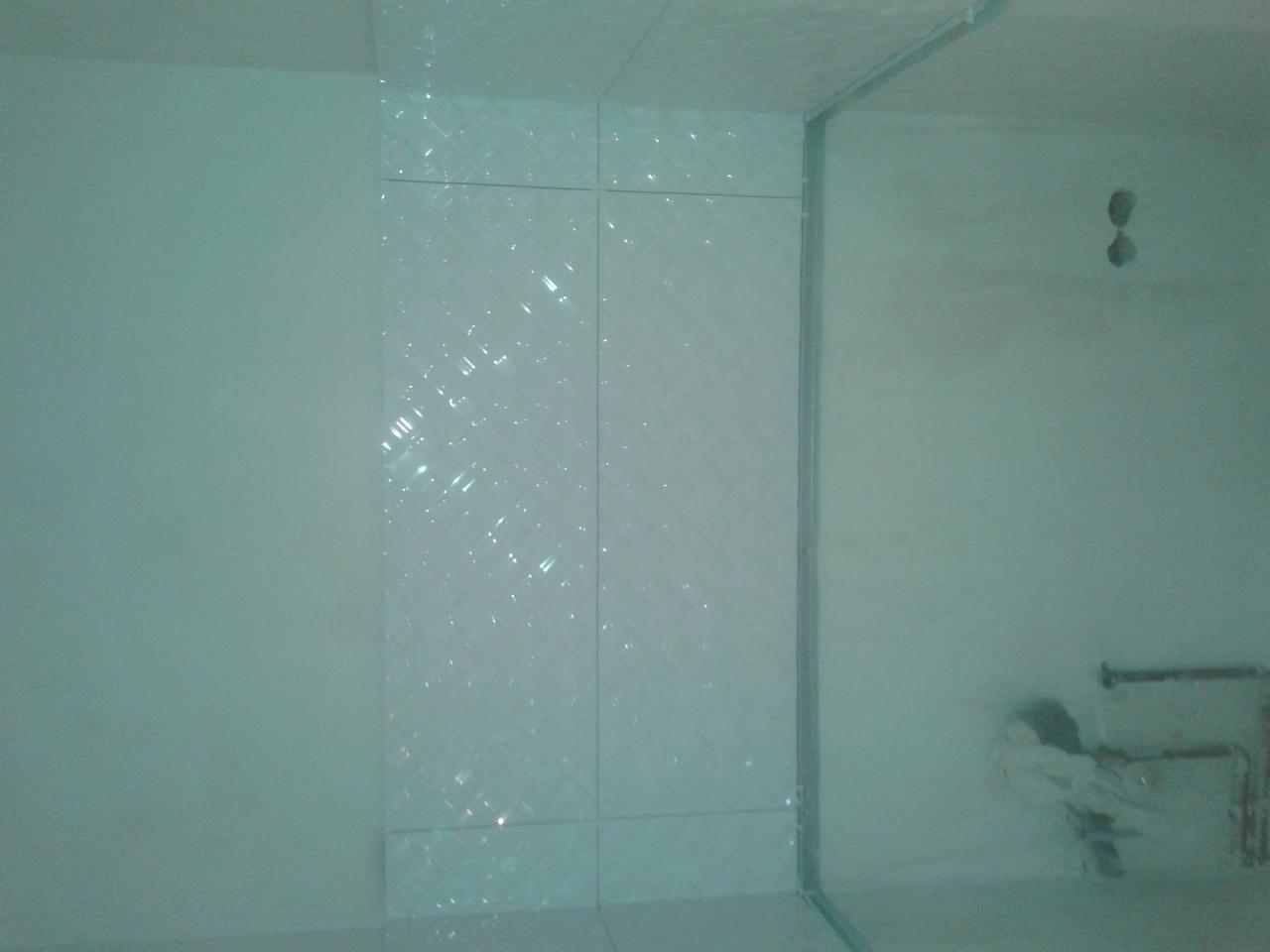 Wykończona ściana kabiny prysznicowej z białymi, połyskującymi płytkami o delikatnym wzorze. Widoczne fragmenty instalacji wodnej i otwory w ścianie.