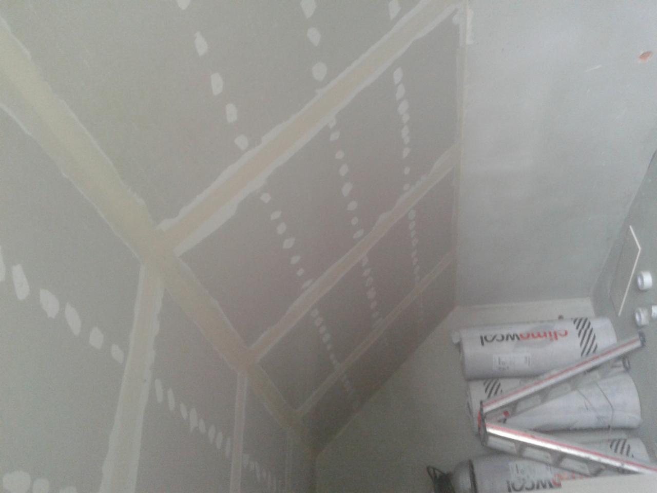 Poddasze po montażu płyt karton-gips, widoczne łączenia wzmocnione taśmą. U dołu leżą rolki wełny izolacyjnej, przygotowane do ocieplenia.