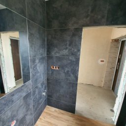 Jony Usługi Remontowo-Budowlane - Łazienka w trakcie wykańczania: ściany wyłożone ciemnoszarą płytką imitującą kamień, drewnopodobna podłoga, widoczne niedokończone instalacje elektryczne i hydrauliczne, surowy stan wykończenia...