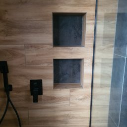 Jony Usługi Remontowo-Budowlane - Kabina prysznicowa z drewnopodobnymi płytkami, wbudowanymi półkami w kolorze grafitowym, czarną armaturą i szklaną ścianką.