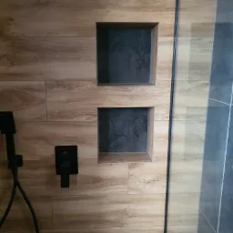 Kabina prysznicowa z drewnopodobnymi płytkami, wbudowanymi półkami w kolorze grafitowym, czarną armaturą i szklaną ścianką.