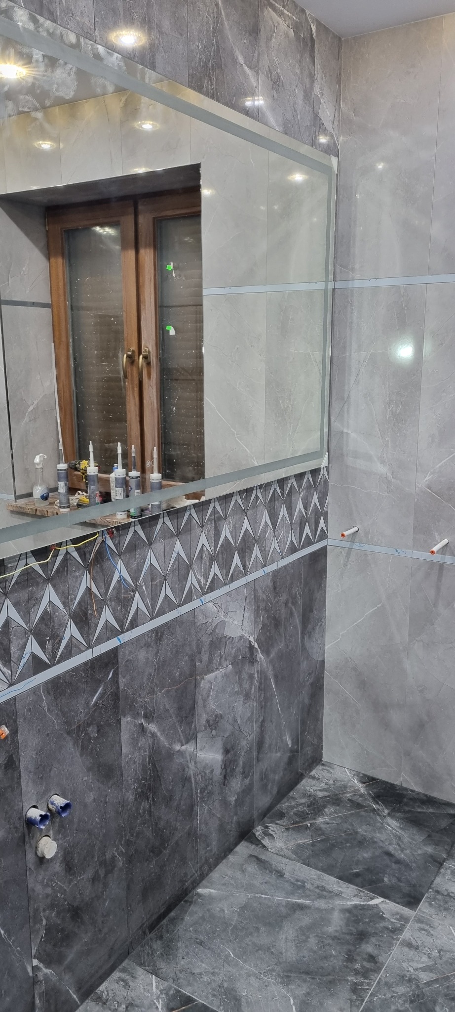 Łazienka w trakcie wykańczania: szare płytki imitujące marmur na ścianach i podłodze, dekoracyjny pas z mozaiki geometrycznej, częściowo zamontowane lustro z widocznym odbiciem okna i narzędzi...