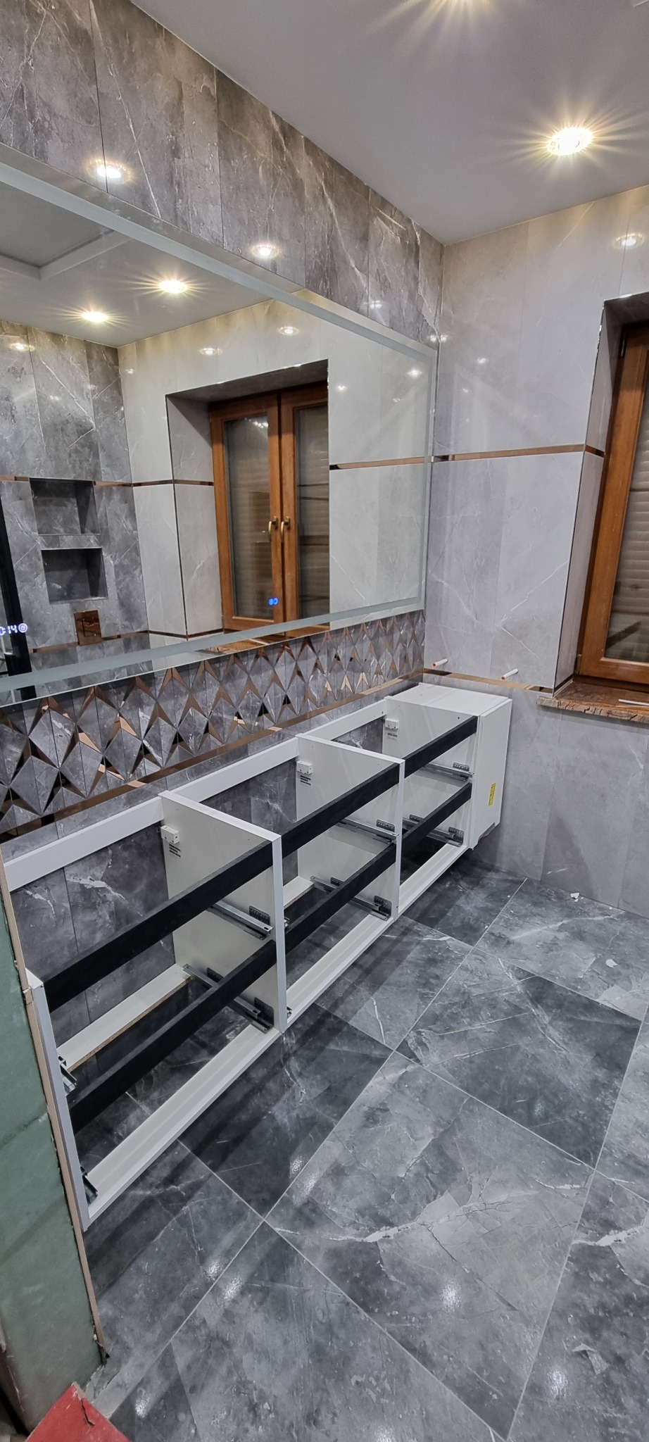 Łazienka w trakcie montażu szafki pod umywalkę, widoczne szuflady bez frontów, marmurowe płytki na ścianach i podłodze, lustro z dekoracyjnym wzorem.