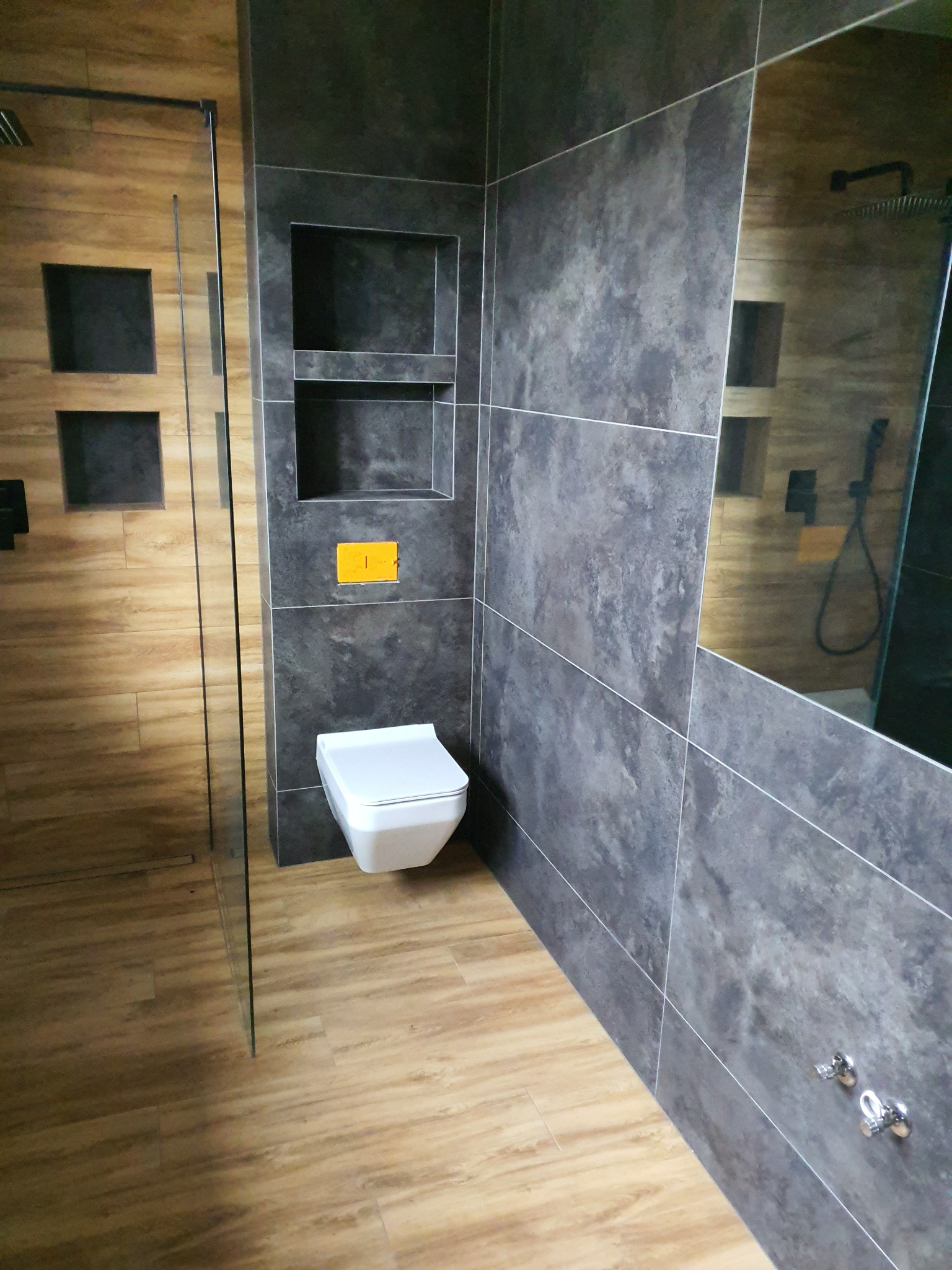 Nowoczesna łazienka z drewnopodobnymi płytkami, szarą ścianą z wnękami i podwieszaną toaletą z żółtym przyciskiem spłukiwania