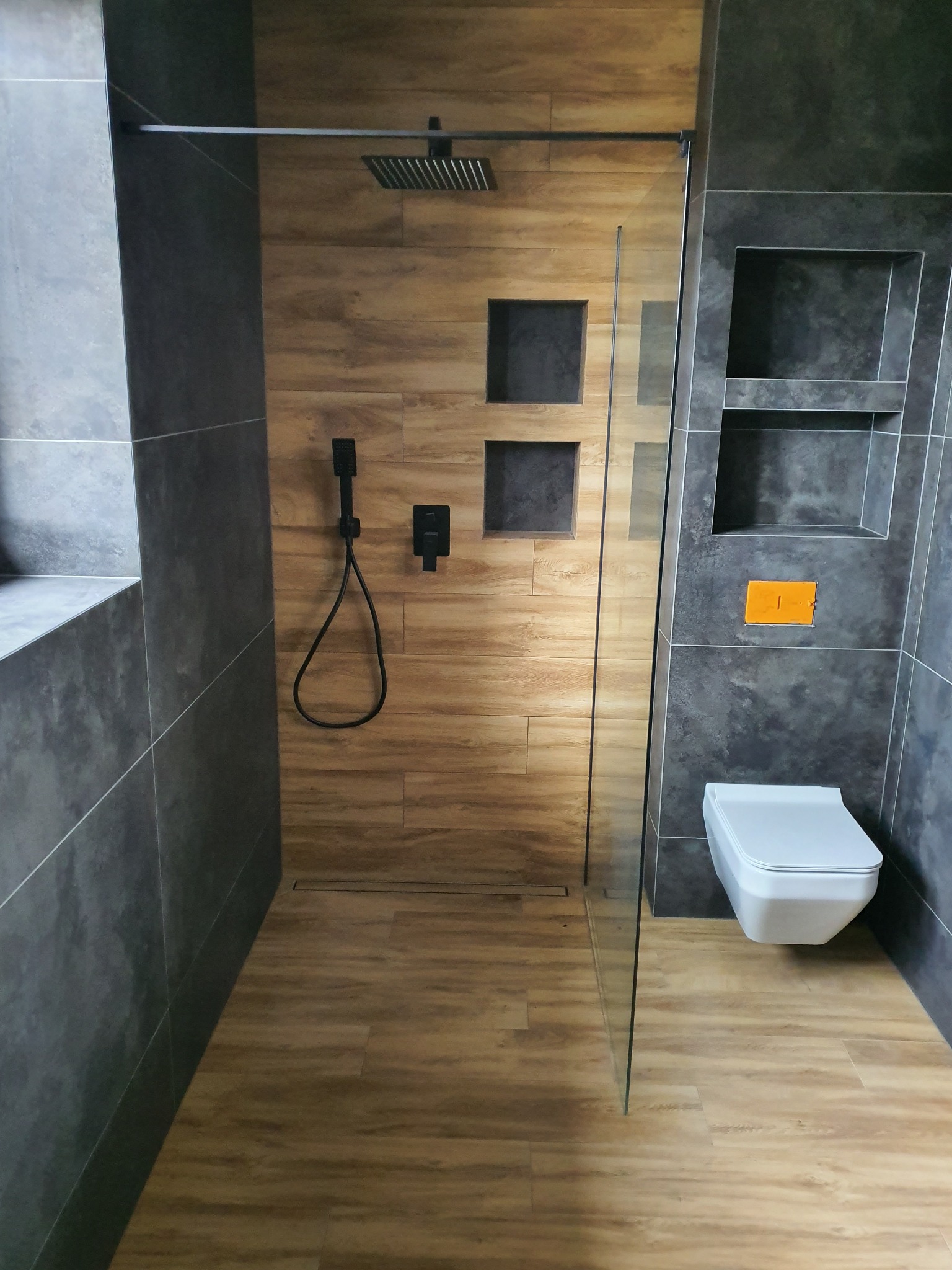 Nowoczesna łazienka z kabiną prysznicową typu walk-in, wykończona płytkami w odcieniach szarości i drewna, z czarną armaturą i podtynkową spłuczką.