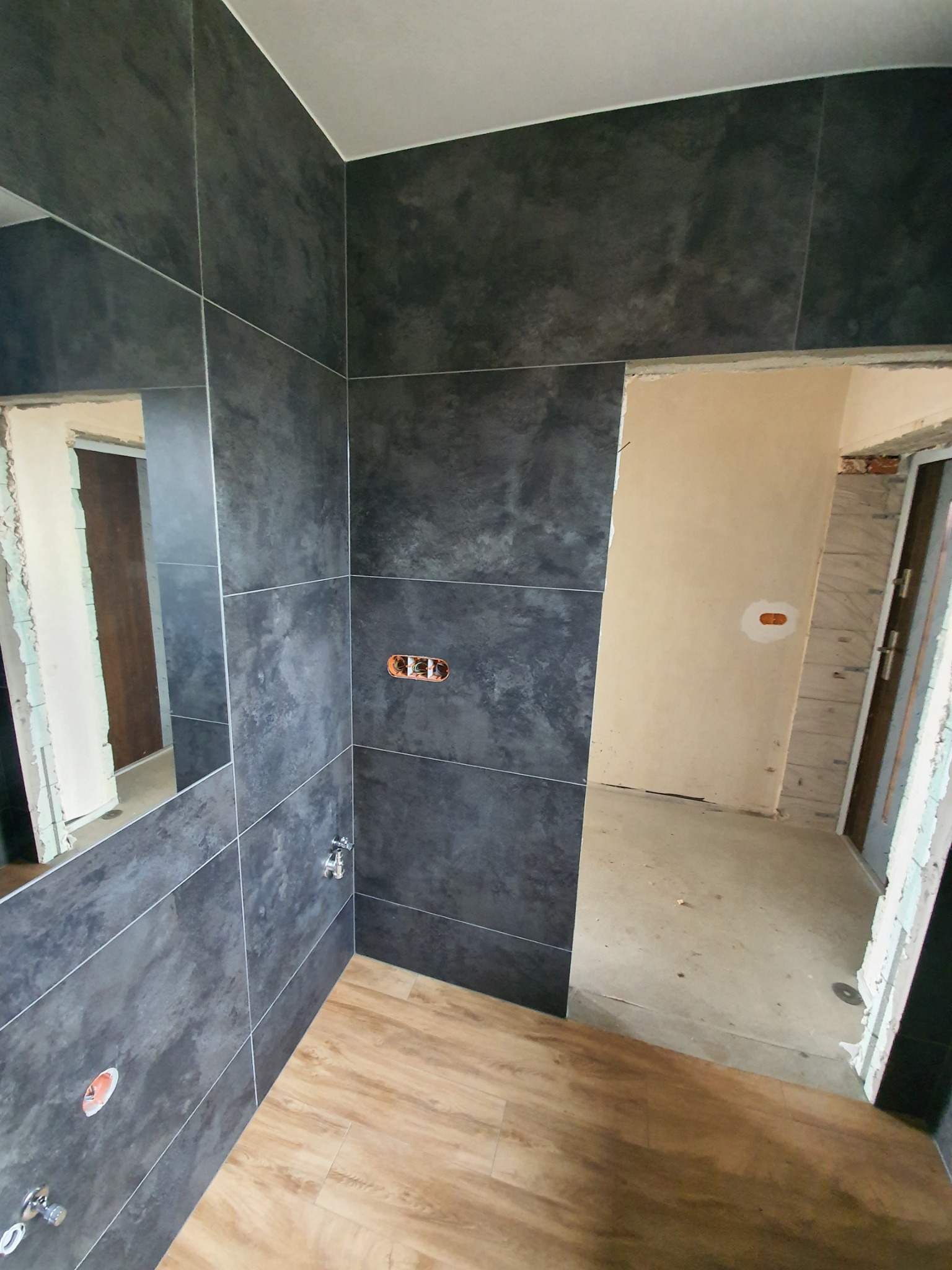 Łazienka w trakcie wykańczania: ściany wyłożone ciemnoszarą płytką imitującą kamień, drewnopodobna podłoga, widoczne niedokończone instalacje elektryczne i hydrauliczne, surowy stan wykończenia...