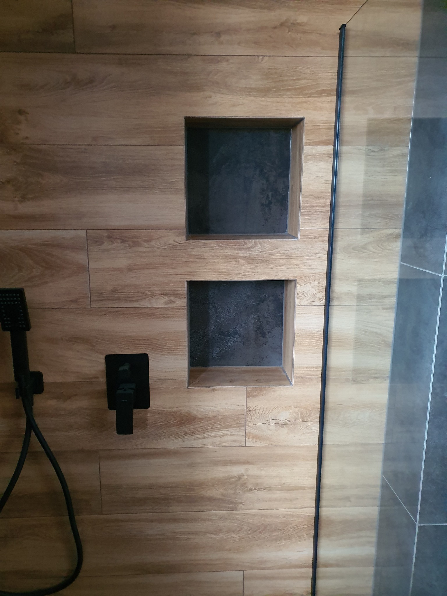 Kabina prysznicowa z drewnopodobnymi płytkami, wbudowanymi półkami w kolorze grafitowym, czarną armaturą i szklaną ścianką.