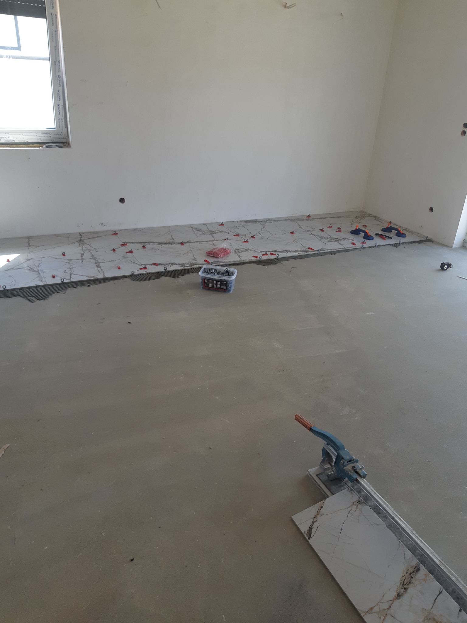 Układanie płytek podłogowych z użyciem poziomujących klipsów i szczypiec, obok leży gilotyna do cięcia płytek, widoczny fragment ściany z gniazdkiem.