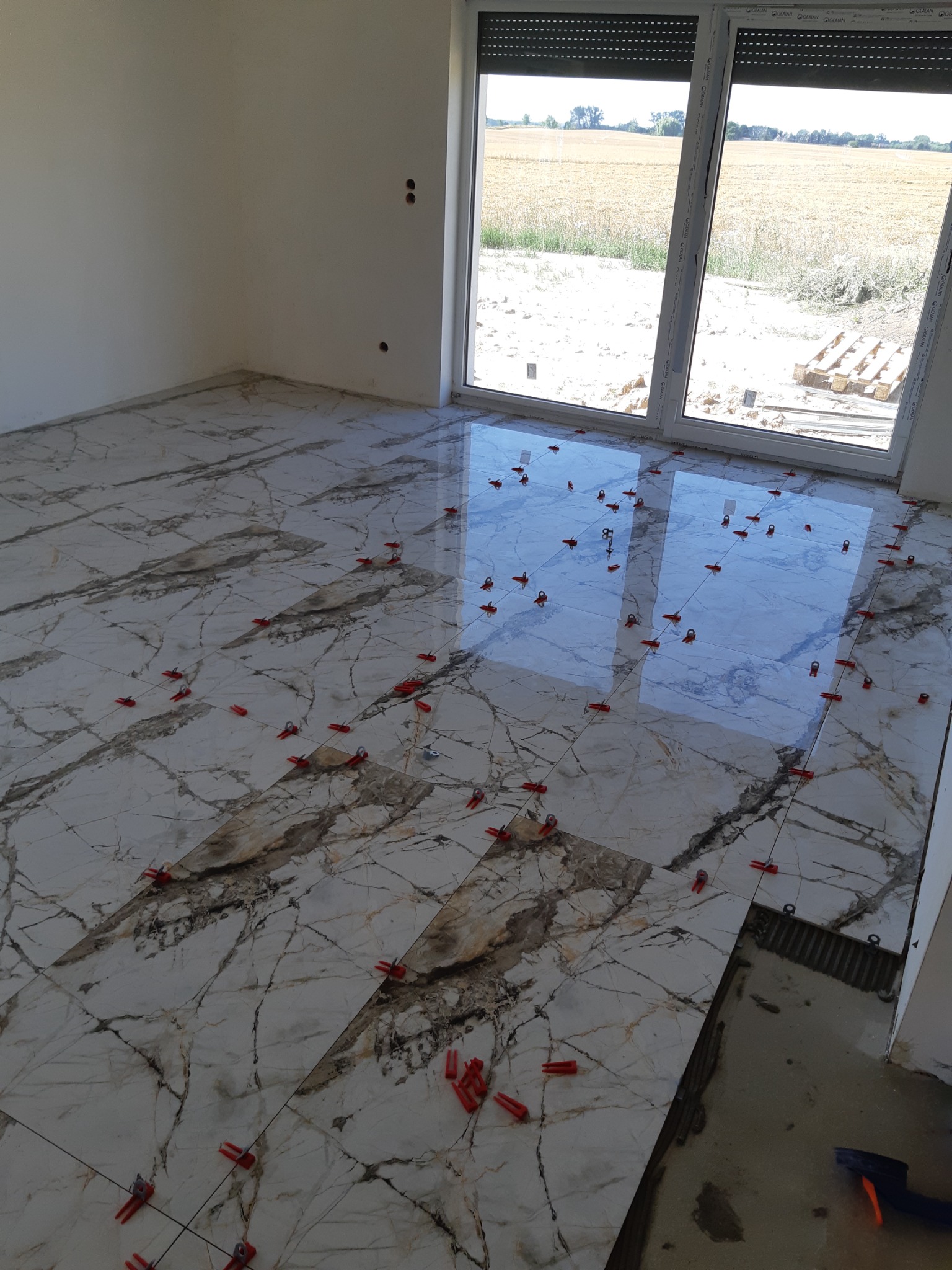 Układanie płytek imitujących marmur na podłodze w jasnym pomieszczeniu z dużym oknem; widoczne krzyżyki dystansowe i klej