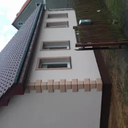 Jasna elewacja domu jednorodzinnego z brązowymi elementami dekoracyjnymi wokół okien i u dołu ściany, widoczny dach z brązowej dachówki i fragment drewnianego ogrodzenia.